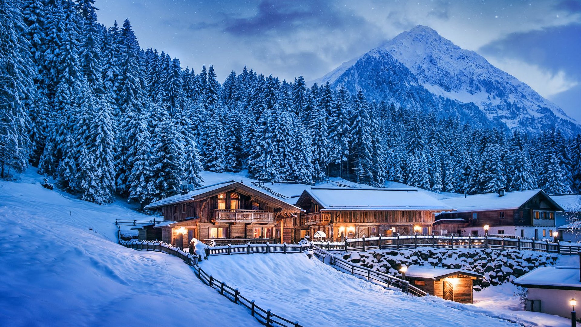 Verschneite Berghütte mit beleuchteten Fenstern und Tannenwald bei Dämmerung